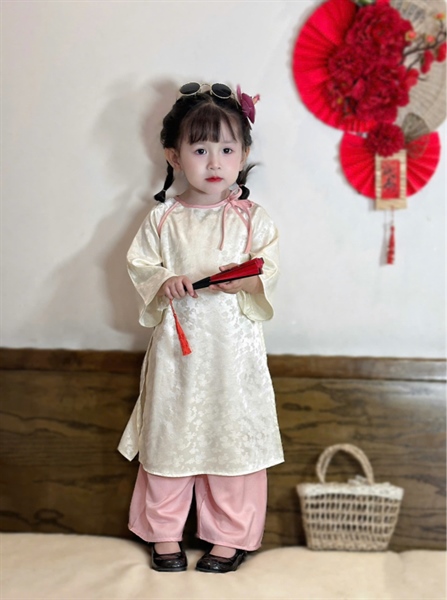 Áo dài bé gái thiết kế cổ tròn màu trắng kem nhạt đính nơ viền tay lửng suông phối quần lụa hồng nude